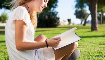 On a bright summer's day, a smiling young woman wearing a summer dress sits cross-legged on a green lawn in a park with a tree in the background reading a book to boost her English Language skills.