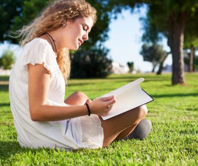 On a bright summer's day, a smiling young woman wearing a summer dress sits cross-legged on a green lawn in a park with a tree in the background reading a book to boost her English Language skills.